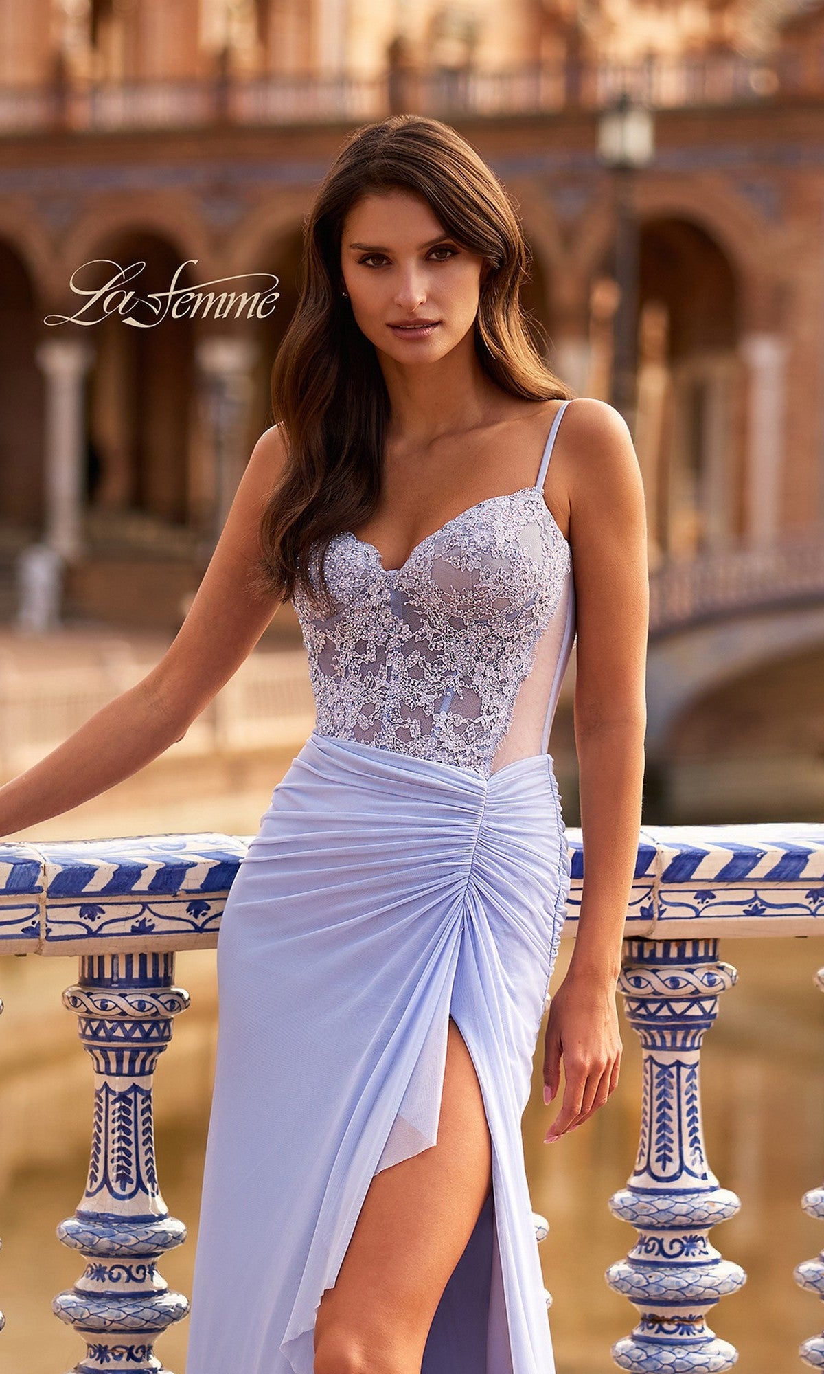A woman poses outdoors by a decorative railing in the Long Prom Dress: La Femme 33661, a light purple gown with a beaded lace corset and thigh-high slit, with arches and water in the background.